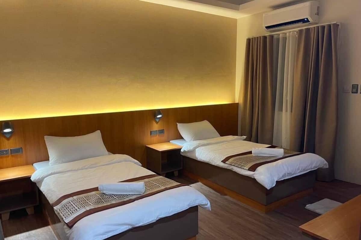 Une chambre d’hôtel avec deux lits, des têtes de lit en bois, des tables de chevet et une grande fenêtre avec des rideaux.