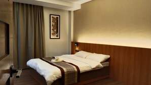 Une chambre d’hôtel avec un lit, une tête de lit, une table de chevet, une lampe, un tableau encadré et une fenêtre avec des rideaux.