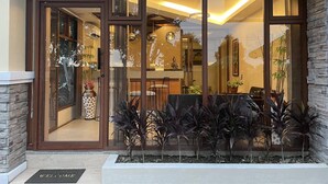 L’entrée de l’hôtel est agrémentée d’un paillasson où est inscrit « Bienvenue », d’un comptoir de réception et d’un espace salon visible à travers les portes vitrées.