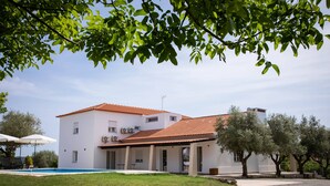 Un bâtiment blanc à deux étages, surmonté d’un toit recouvert de tuiles rouges, avec une piscine et des oliviers au premier plan.