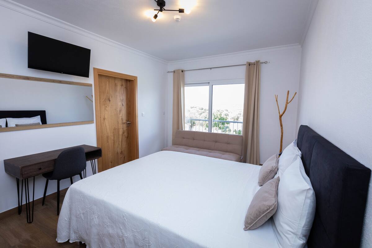 Ein Schlafzimmer mit Bett, Schreibtisch, Stuhl, Fernseher und einem Fenster mit Vorhängen.