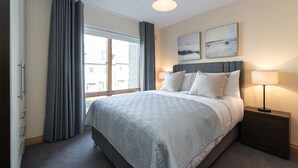 A bedroom with a large bed, a nightstand, a window with curtains, and two wall-mounted artworks. - Charming Leopardstown Townhouse (Dublin)