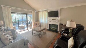 A living room with a sofa, armchair, ottoman, fireplace, TV, and a view through large windows.