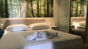 Cama bem arrumada com lençóis e toalhas brancos, mesa de cabeceira com abajur e um espelho grande refletindo o quarto.