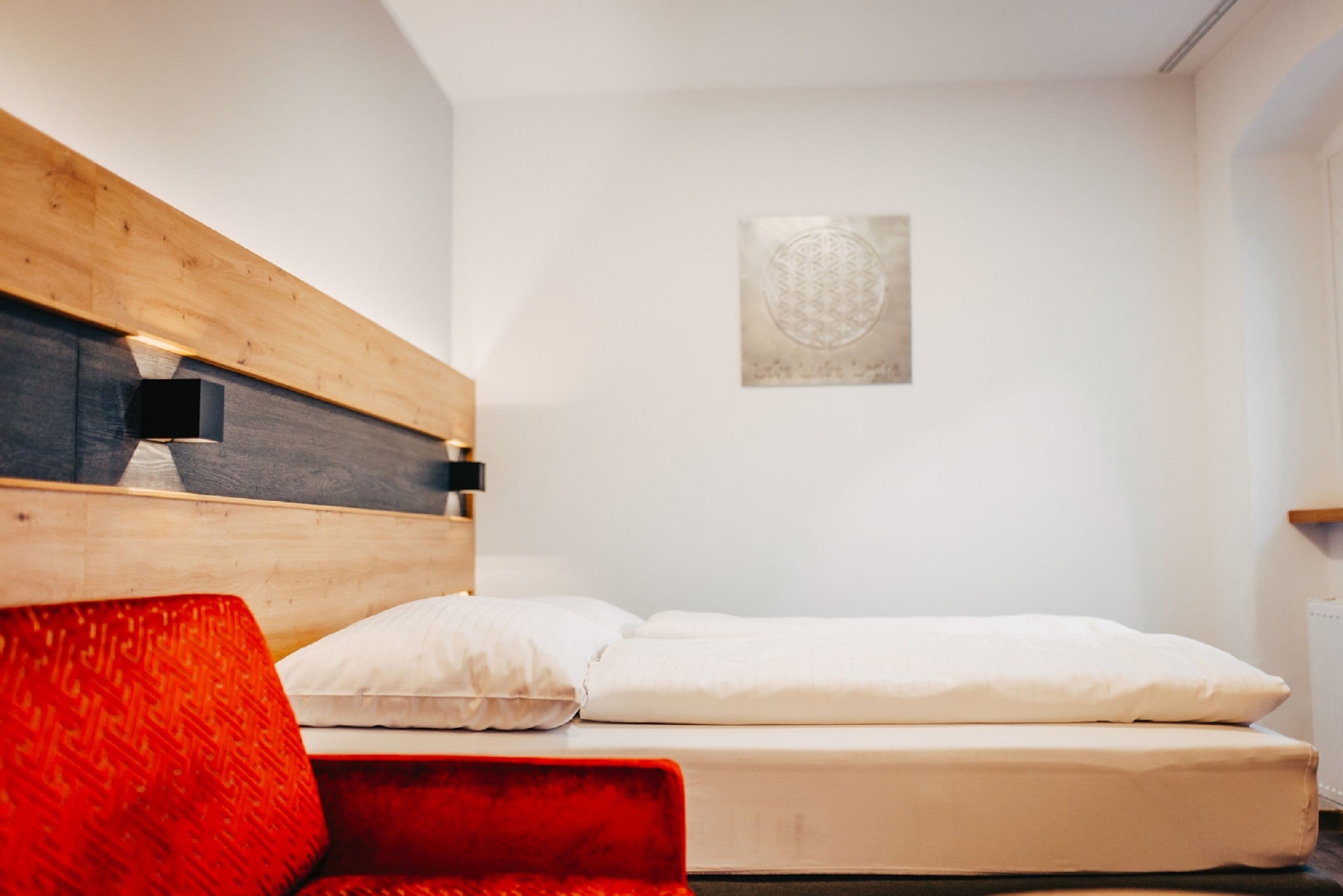 A modern bedroom with a wooden headboard, a red chair, a bed with white bedding, and a wall-mounted artwork.
