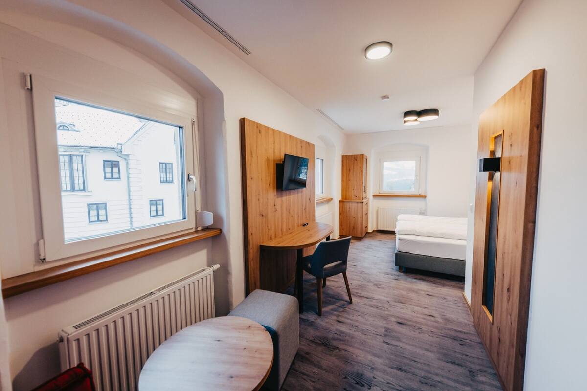 Habitación compacta con cama, una mesita, una silla, televisor montado en la pared y ventana con vistas a edificios.