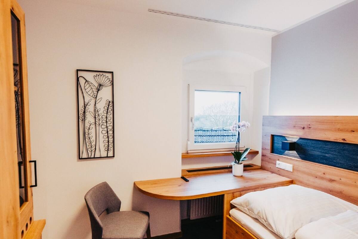 Una habitación pequeña con cama, escritorio, silla, ventana y una obra de arte en la pared.