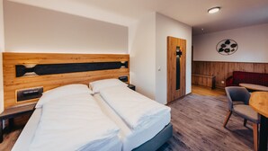 A hotel room with a wooden headboard, a bed with white linens, a small bedside table, a chair, a round table, and a wall-mounted artwork.