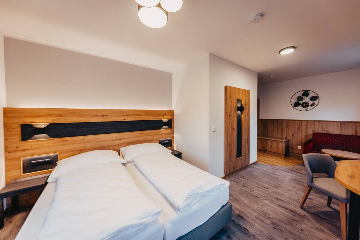 Una habitación de hotel con cabecera de madera, cama con sábanas blancas, mesita de noche, una silla, una mesa redonda y una obra de arte colgada en la pared.