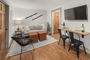 A modern living room with a sofa, ottomans, a coffee table, a TV, and a dining area with chairs and a table. - Stowe Dual Condo | 4BR Mountain & River Retreat (Stowe)