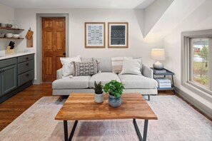 A living room with a sofa, coffee table, and decorative items. There is a wooden door, a window, and a lamp on a side table.