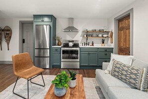 A modern kitchen with teal cabinets, stainless steel appliances, and a wooden dining table with plants. - Stowe Dual Condo | 4BR Mountain & River Retreat (Stowe)