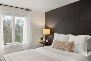 A bedroom with a bed, a bedside table with a lamp, a clock, and a window with curtains. - Stowe Dual Condo | 4BR Mountain & River Retreat (Stowe)