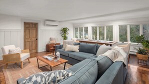 A modern living room with a grey sofa, wooden coffee table, and a large window offering a view of trees. - 4BR Vermont Condo | Stowe Mountain & River Access (Stowe)