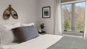 Ein Schlafzimmer mit einem Bett, Kissen, einem Nachttisch mit Pflanze und Uhr, einem gerahmten Bild und einem Fenster mit Vorhängen.