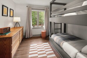 Une chambre avec un lit superposé, une commode en bois, une lampe, une fenêtre avec des rideaux et un tapis à carreaux.