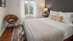 Un dormitorio ordenado con una cama grande, una mesita de noche con lámpara, una silla y vistas al exterior a través de una ventana.