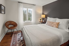 A neatly arranged bedroom with a large bed, a side table with a lamp, a chair, and a view of the outdoors through a window. - Vermont 2BR | Stowe Slopes & Swimming Holes (Stowe)