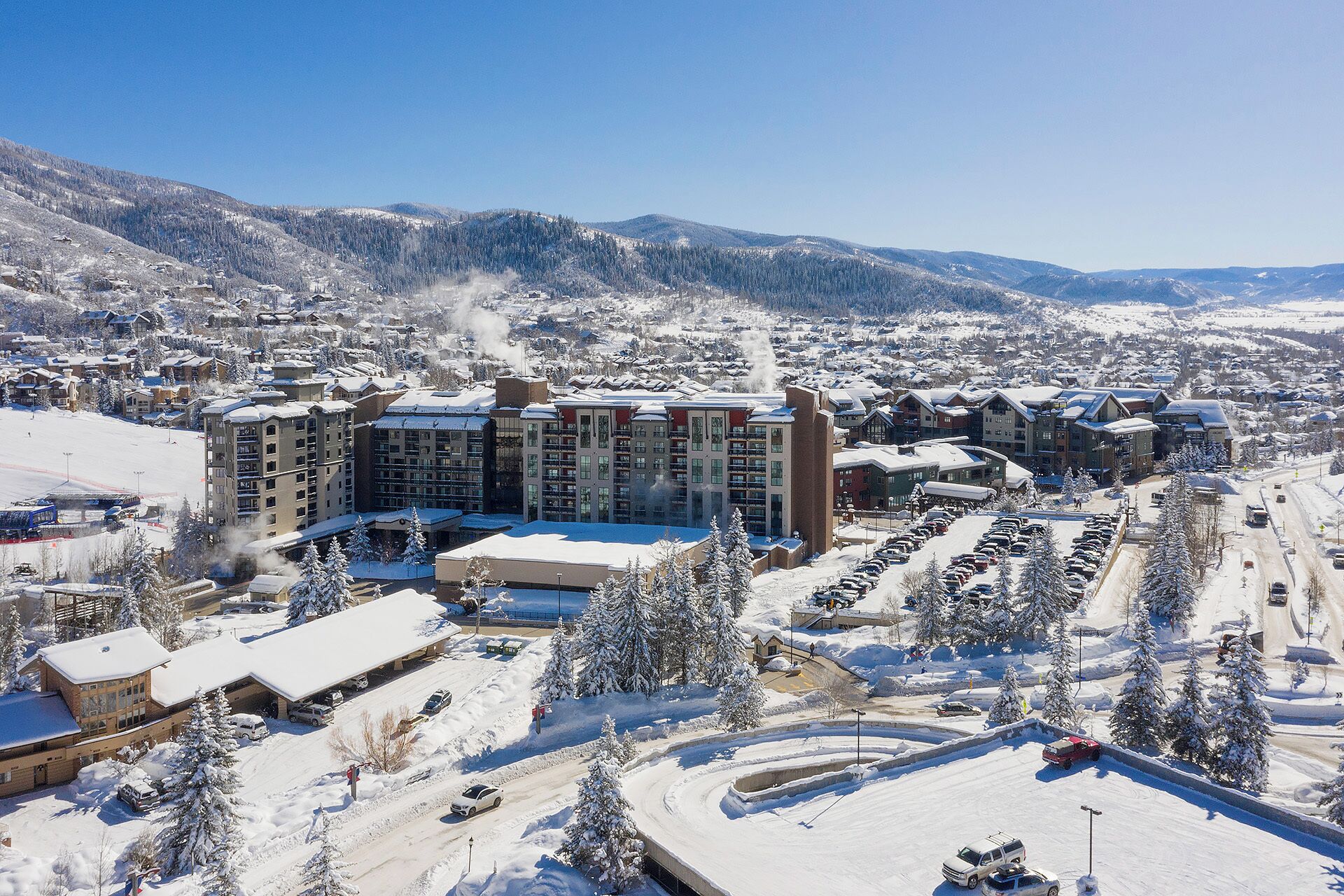 Una località turistica innevata con diversi edifici, tra cui un grande albergo con balconi, circondata da alberi carichi di neve e uno scenario montuoso.