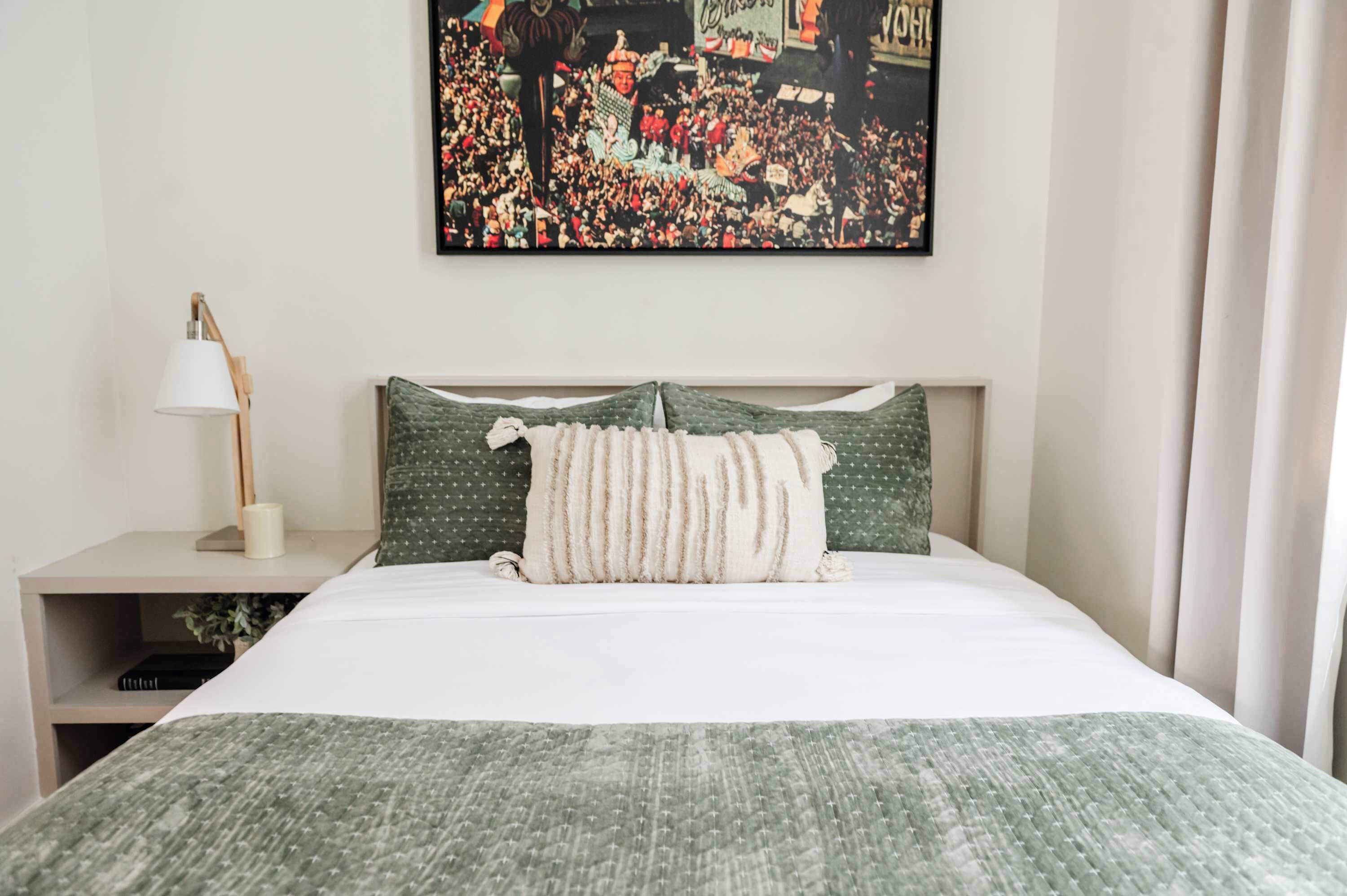 A neatly made bed with a green and white patterned bedspread, a pillow with a similar pattern, and a framed picture of a crowded event on the wall.