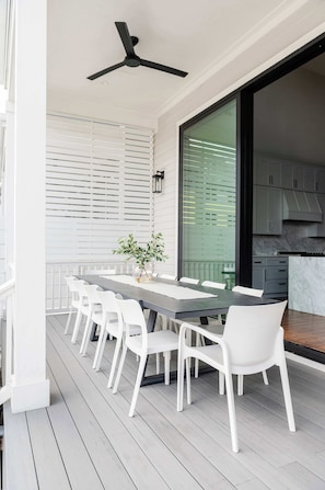 A modern outdoor dining area with a long table and white chairs, a ceiling fan, and a view of a kitchen through a glass door. - The 1300 | Designer 7 bedroom, Heated Pool (New Orleans)