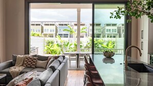 A modern living room with a sofa, a dining table, and chairs. A large sliding glass door leads to an outdoor balcony with a view of other buildings and greenery. - The 1300 | 7 Bedroom Luxe Home, Heated Pool & Spa (New Orleans)
