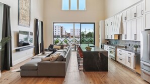 A spacious living area with a long sofa, a kitchen with white cabinets, and a large window overlooking a cityscape. - The 1300 | 7 Bedroom Luxe Home, Heated Pool & Spa (New Orleans)