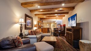 A cozy living room with a sofa, ottoman, and coffee table, wooden beams on the ceiling, and a television on the right.