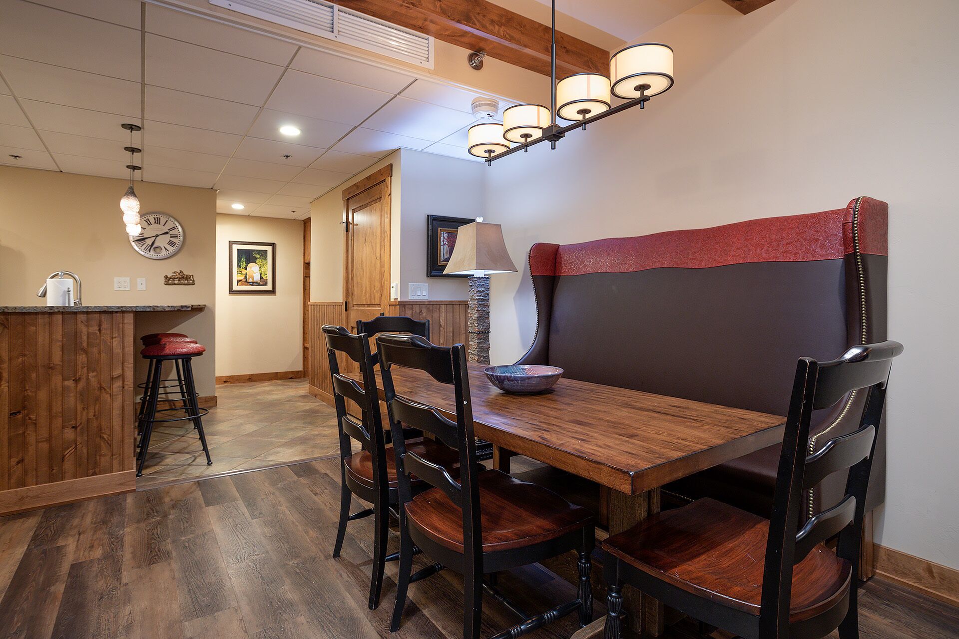 Un comedor con una mesa de madera, cuatro sillas, un banco con cojín rojo, un reloj de pared y una lámpara colgante.