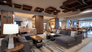 Un hall d’hôtel moderne avec un canapé d’angle, des fauteuils, une table basse et un téléviseur. On y trouve également des étagères en bois et un présentoir à eau.