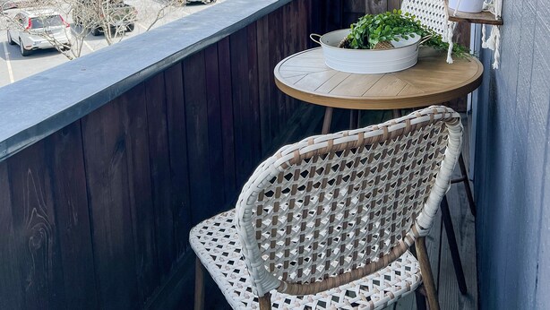 En balkon med gelænder i træ, en fletstol, et lille rundt bord og en potteplante.
