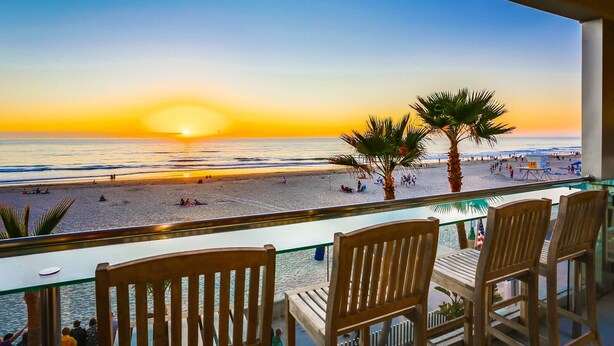 A balcony with wooden chairs overlooking a beach at sunset.