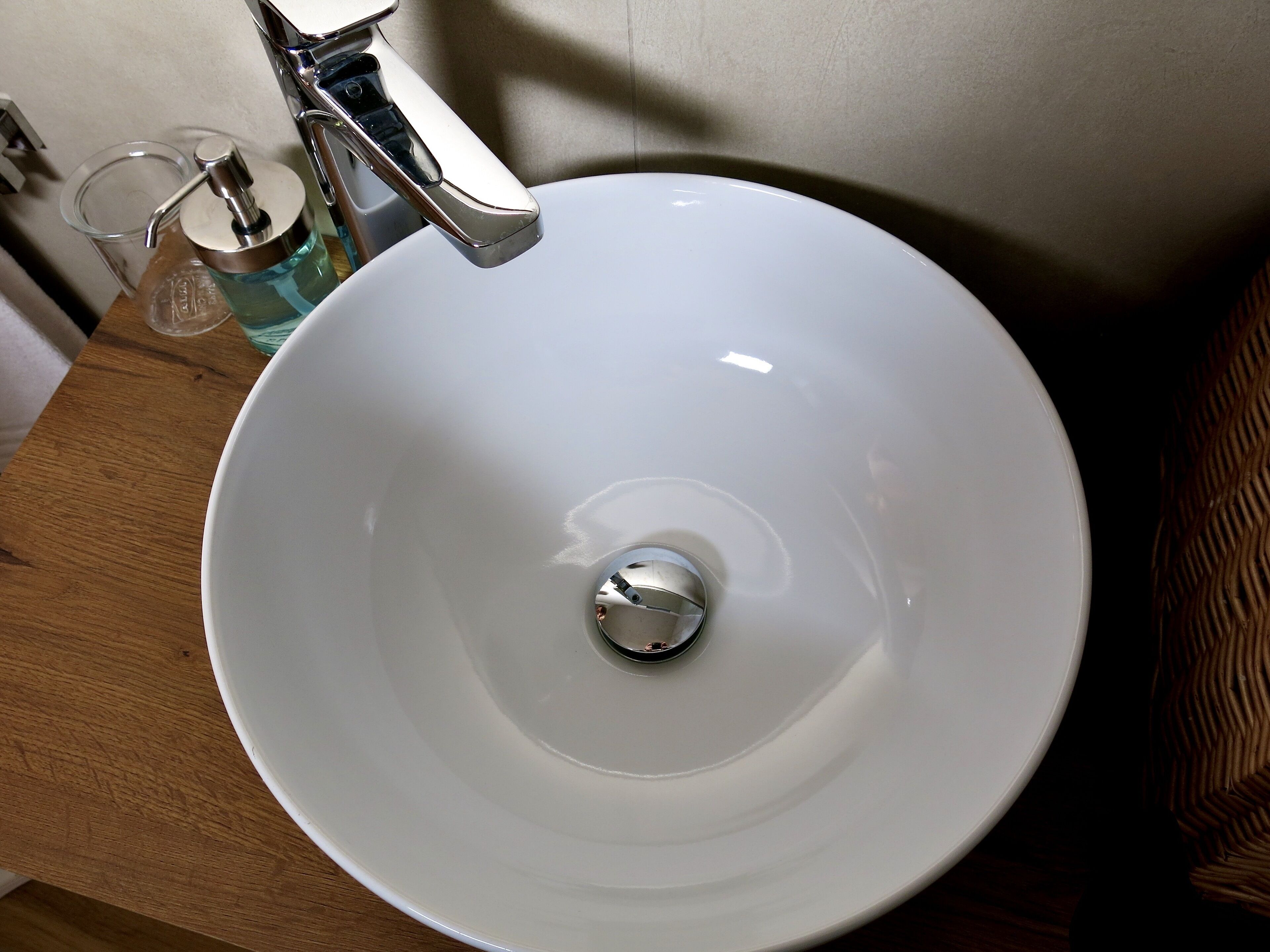 Un évier en céramique blanche, doté d’un robinet chromé, un verre transparent et un flacon de savon, le tout posé sur un comptoir en bois.