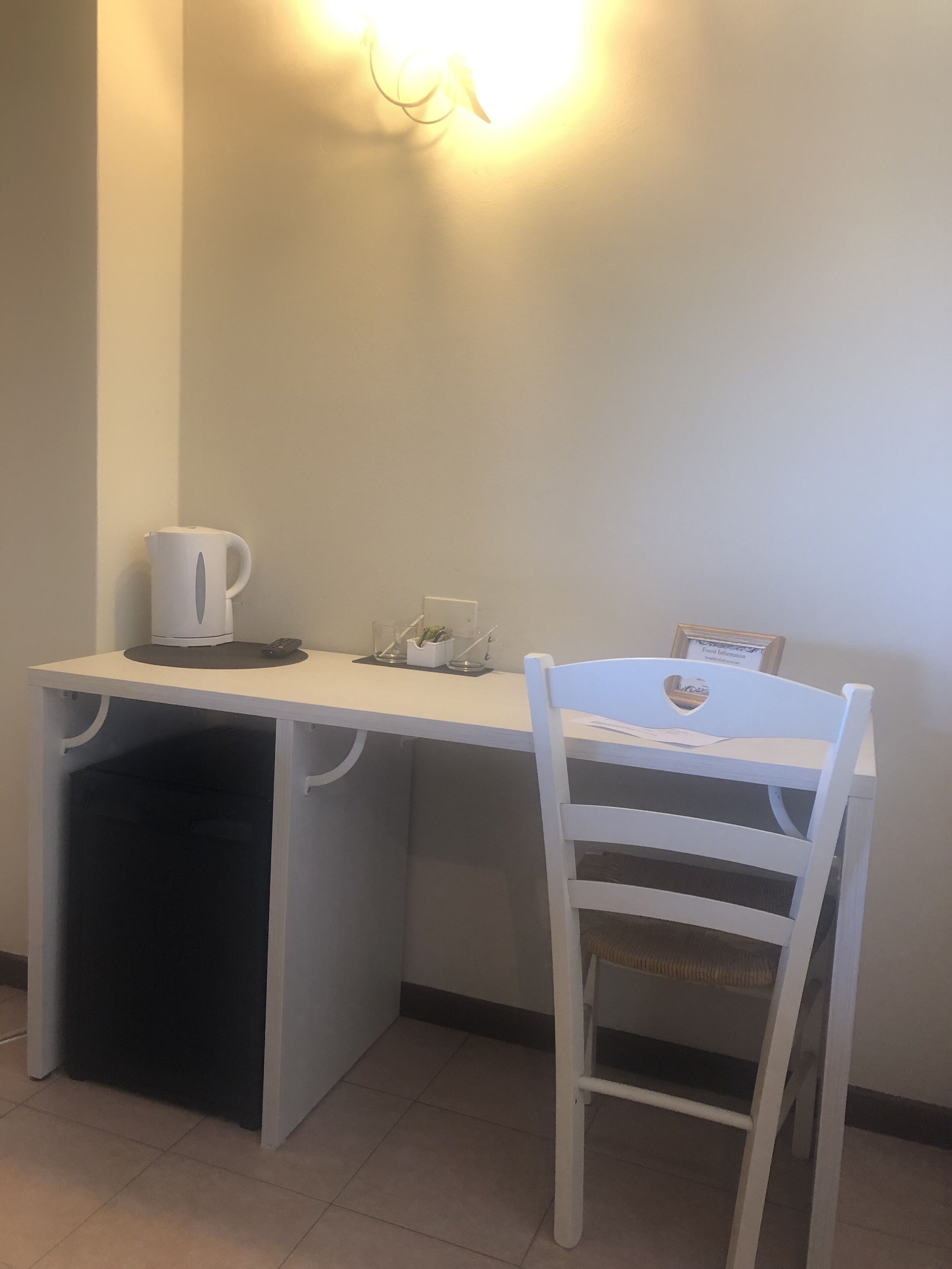 Een klein wit bureau met een kopje en een plantje erop, een witte stoel met een hartvormige uitsparing in de rugleuning, en een wandlamp.