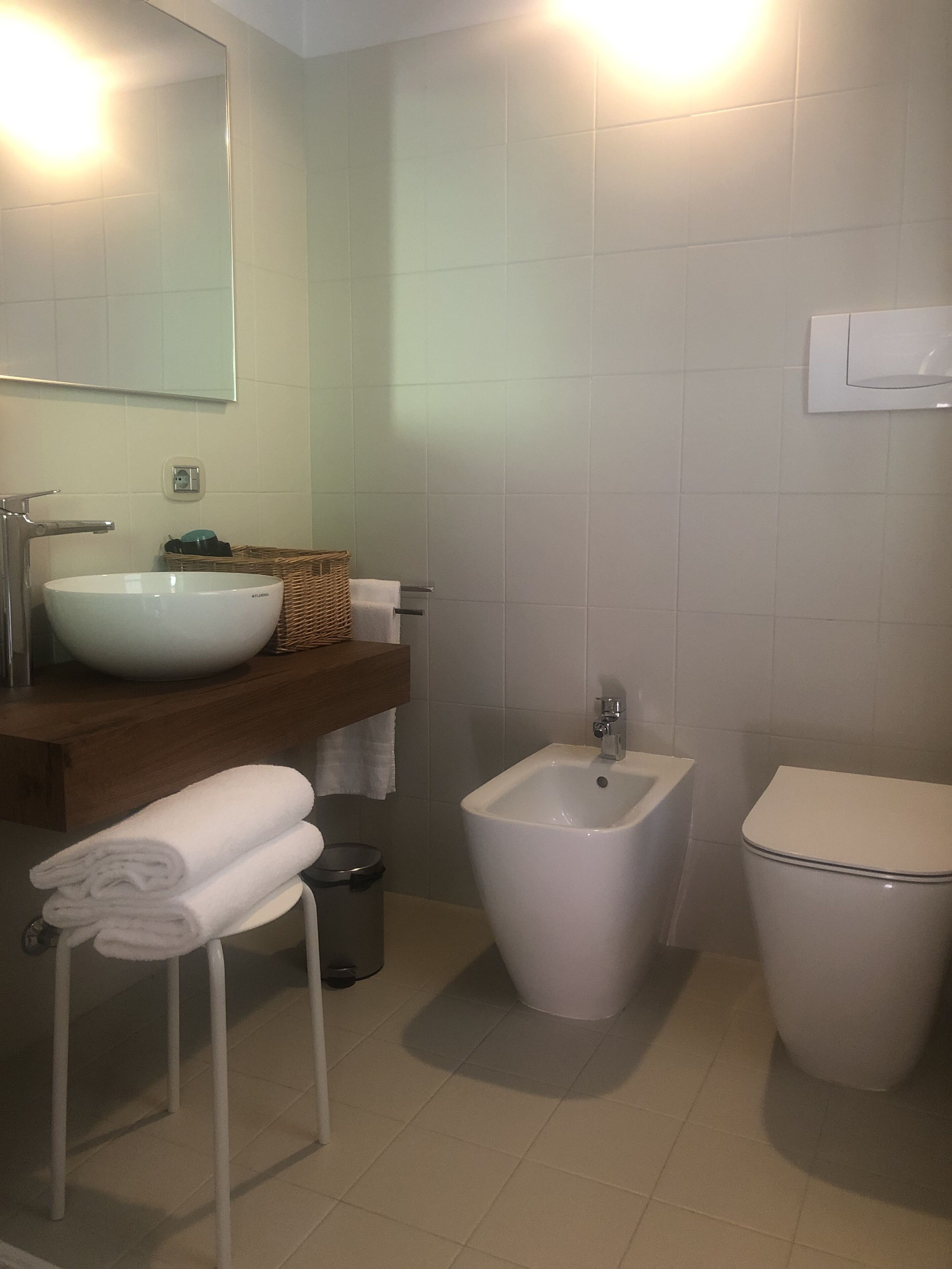 Une salle de bain avec un lavabo blanc, un bidet et des toilettes. On y trouve un porte-serviettes avec des serviettes pliées et un panier en osier posé sur le comptoir.