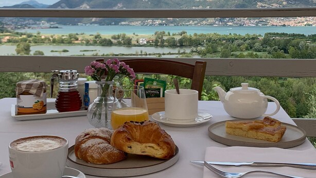 A table set with breakfast items, including croissants, a cup of coffee, and a teapot, overlooking a scenic view of mountains and a lake. - AGRITURISMO FOREST B&B (Iseo)