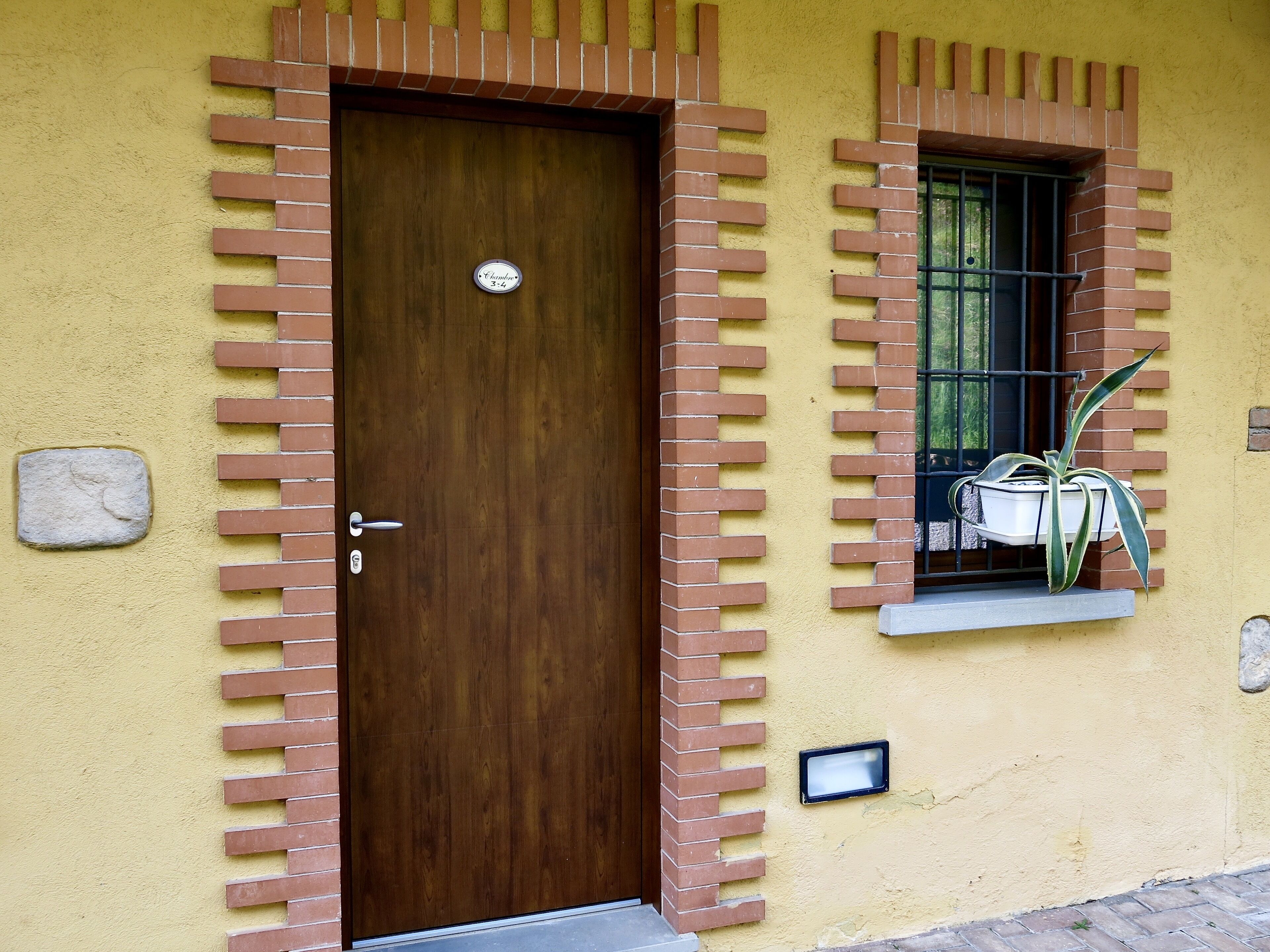 Una puerta de madera con manija metálica, adornada con detalles de ladrillo, un pequeño ventanal con una planta en maceta y una placa de piedra en la pared.
