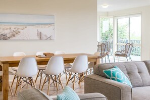 Ein Essbereich mit einem Holztisch und weißen Stühlen, einer Couch mit einem gemusterten Kissen und einem Wandbild mit einer Strandansicht.