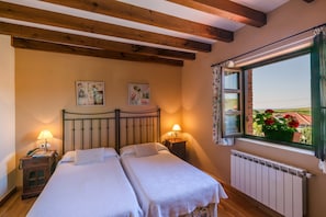 A bedroom with two beds, wooden beams on the ceiling, a window with a view of a garden, and a radiator. - Posada Caborredondo (Alfoz de Lloredo)