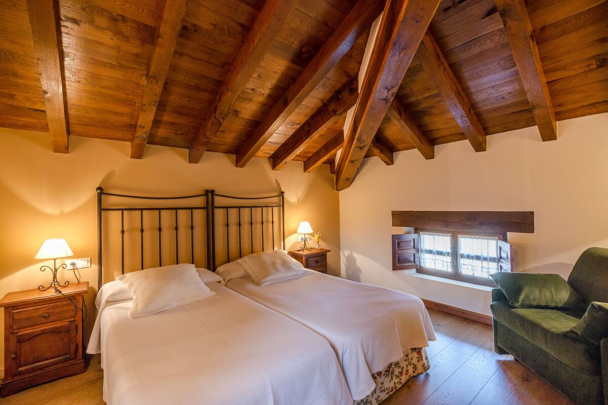 Un dormitorio con techo de madera, una cama grande con ropa de cama blanca, dos mesitas de noche con lámparas, un sillón verde y una ventana con cortinas.