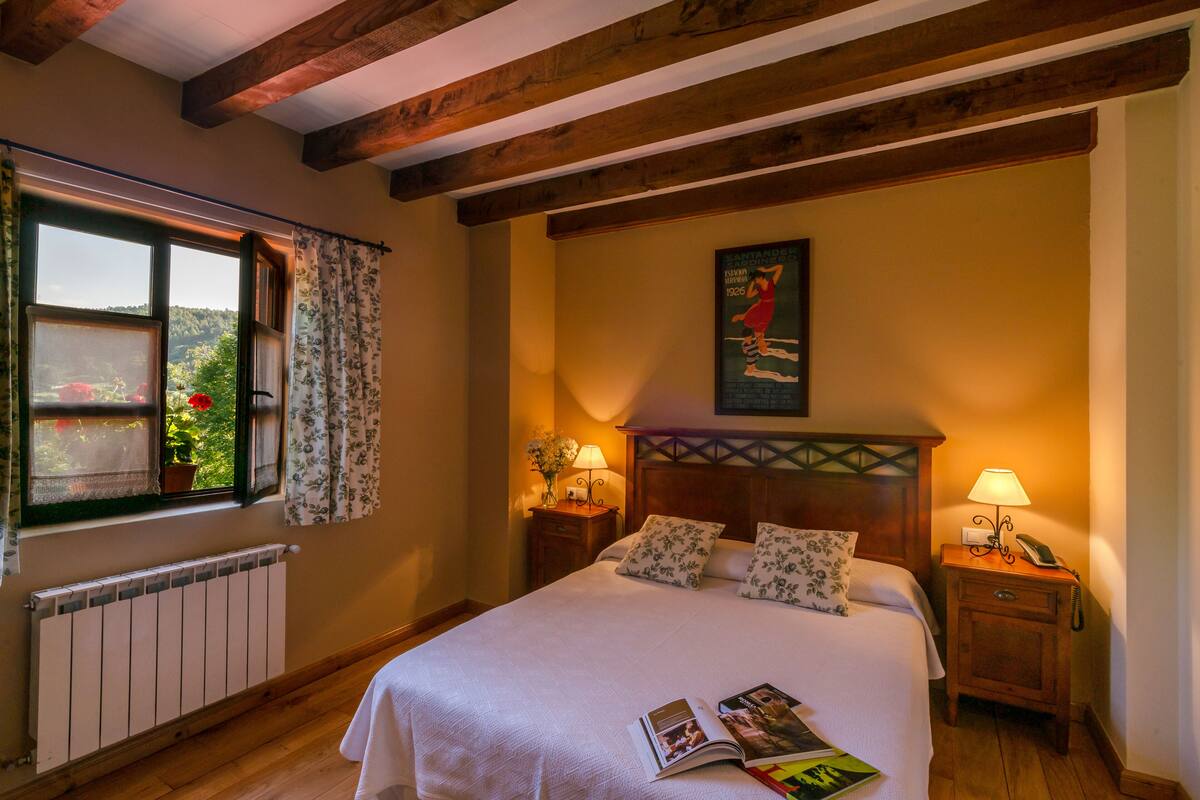 Un dormitorio con una cama grande, dos mesitas de noche con lámparas, un radiador, una ventana con cortinas y un cuadro en la pared.