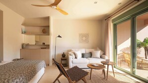 Ein modernes Schlafzimmer mit einem Bett, einem Holzsessel, einem kleinen Tisch und einem Sofa. Es gibt einen Deckenventilator, ein Fenster mit Vorhängen und einen Blick ins Freie.