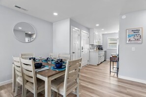 A dining area with a wooden table and chairs, a kitchen in the background, and a framed picture on the wall.