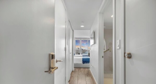 A hallway leading to a bedroom with a bed, a door with a handle, and a wall-mounted light fixture.