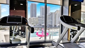 Une salle de sport équipée de tapis de course et dotée d’une grande fenêtre donnant sur une piscine et les bâtiments de la ville.