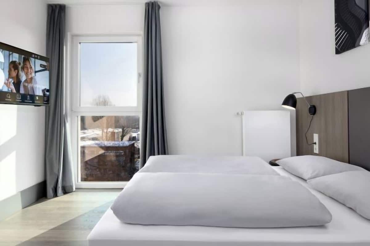 A hotel room with a bed, a TV, a window with curtains, and a view of the outside.