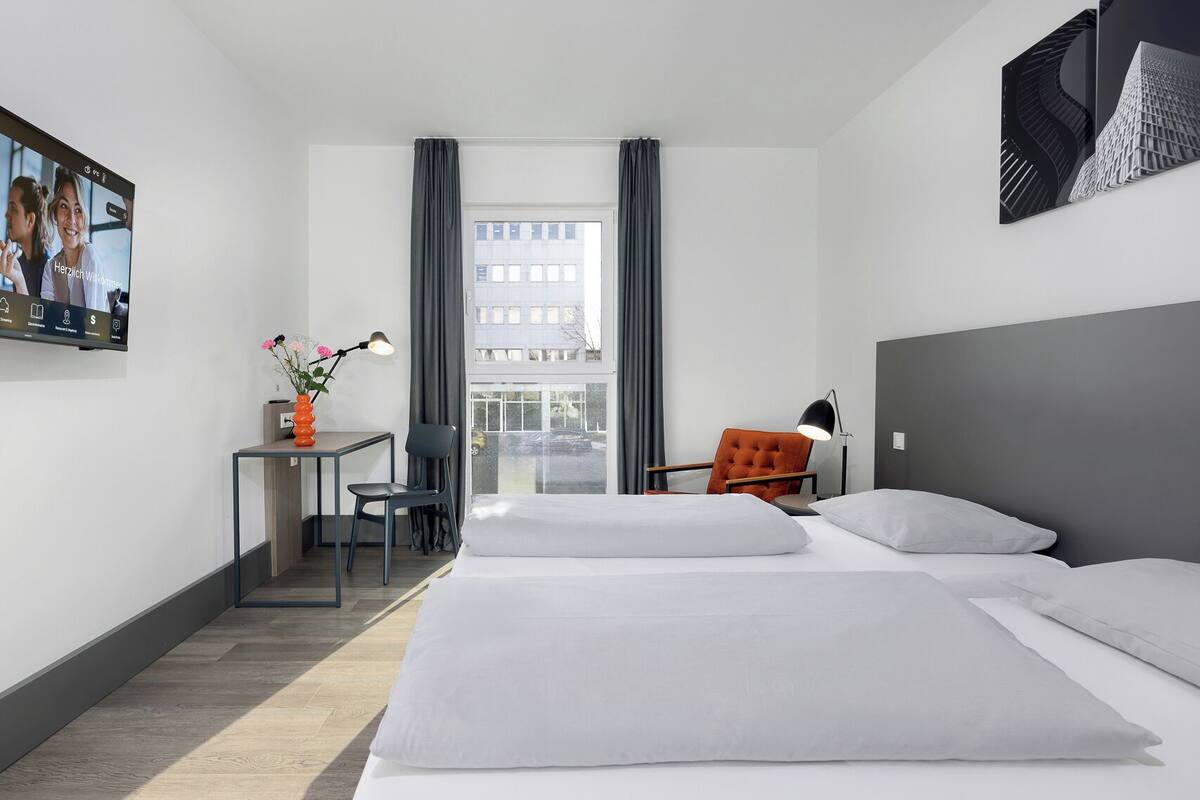 A hotel room with two beds, a desk with a chair, a vase with flowers, and a large window with curtains.