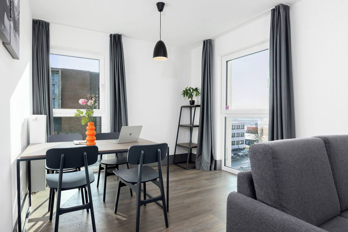 A modern living room with a grey sofa, a wooden dining table with four chairs, a laptop, and a potted plant.