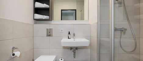 Un baño moderno con inodoro, lavabo y ducha. Hay estantes con toallas y artículos de aseo.