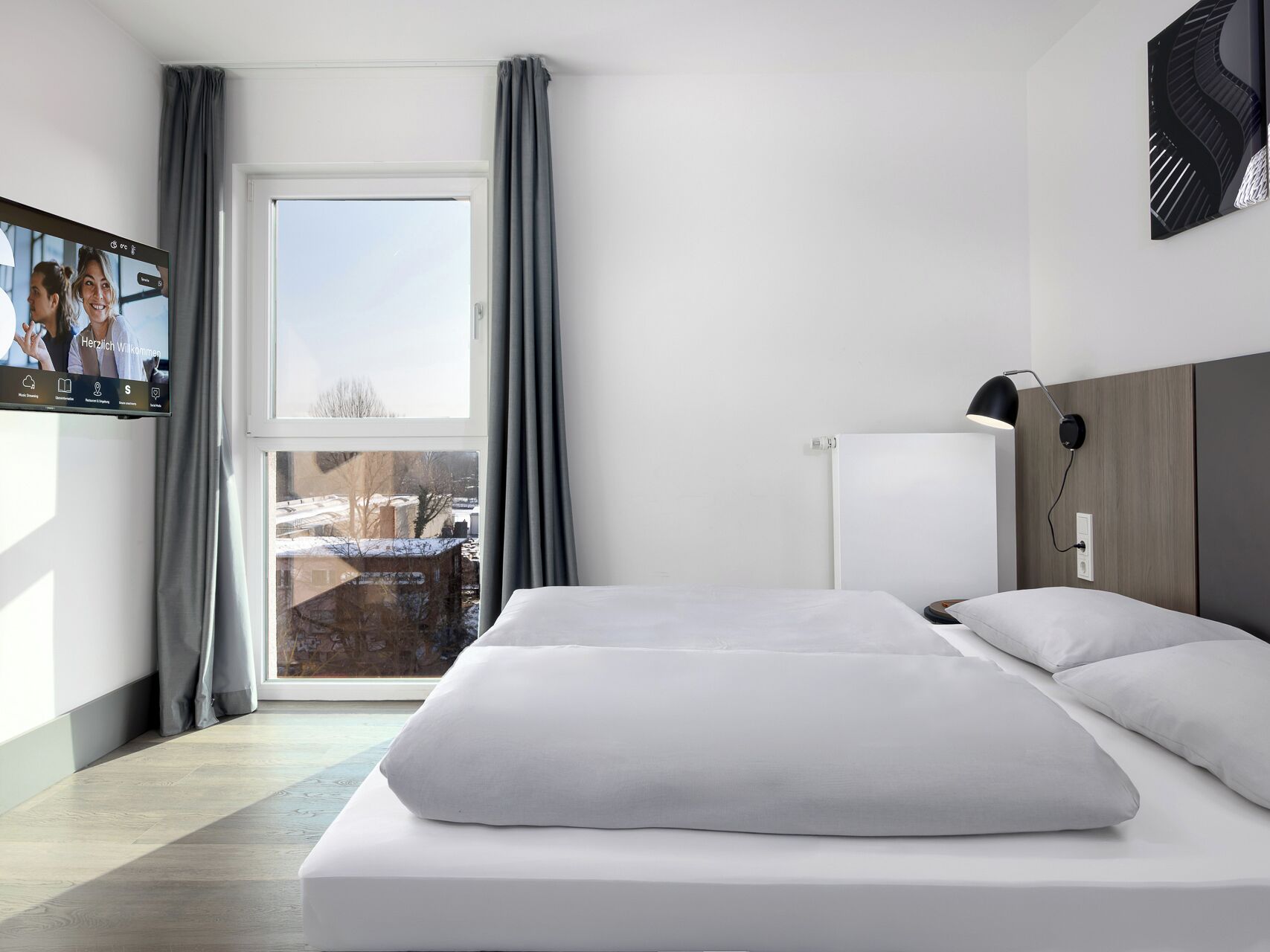 A modern hotel room with a large bed, a flat-screen TV, a desk lamp, and a view of the outside.